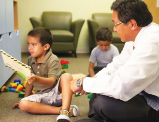 Dad sitting on the floor playing with his young son while other son is in background