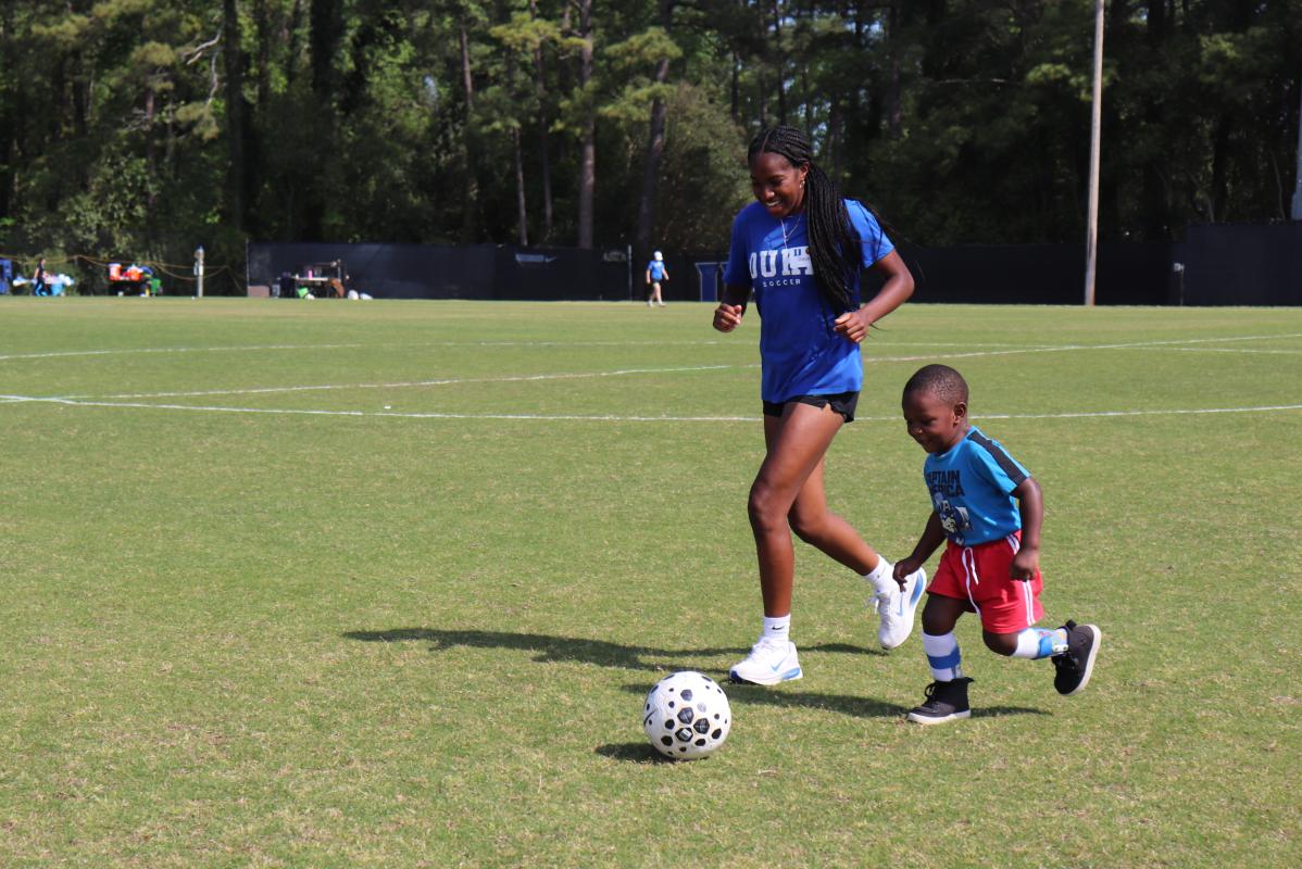 Duke Womens soccer player running with a young boy dribbling a soccer ball