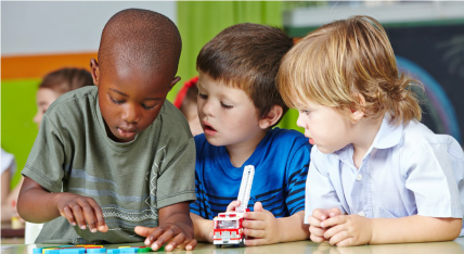 a group of 3 boys about 5 years old look at school work together at a table.
